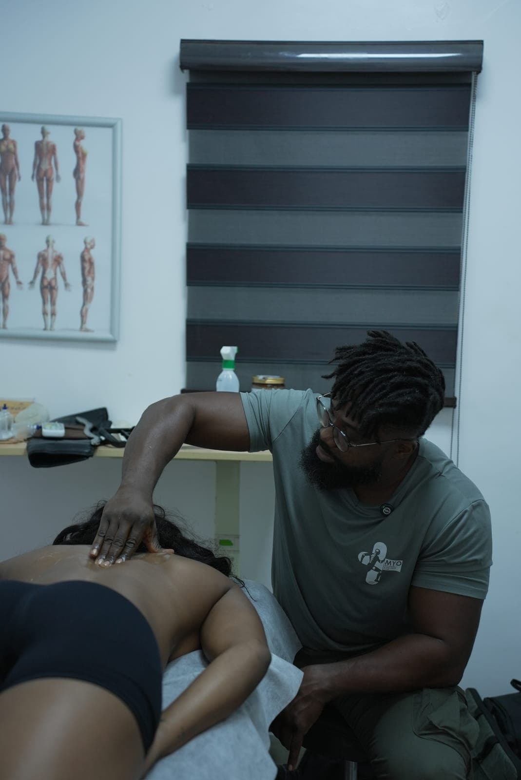 The MYO treatment room: a therapist seated at the couch working on a client's back, the anatomy chart and a striped blind behind