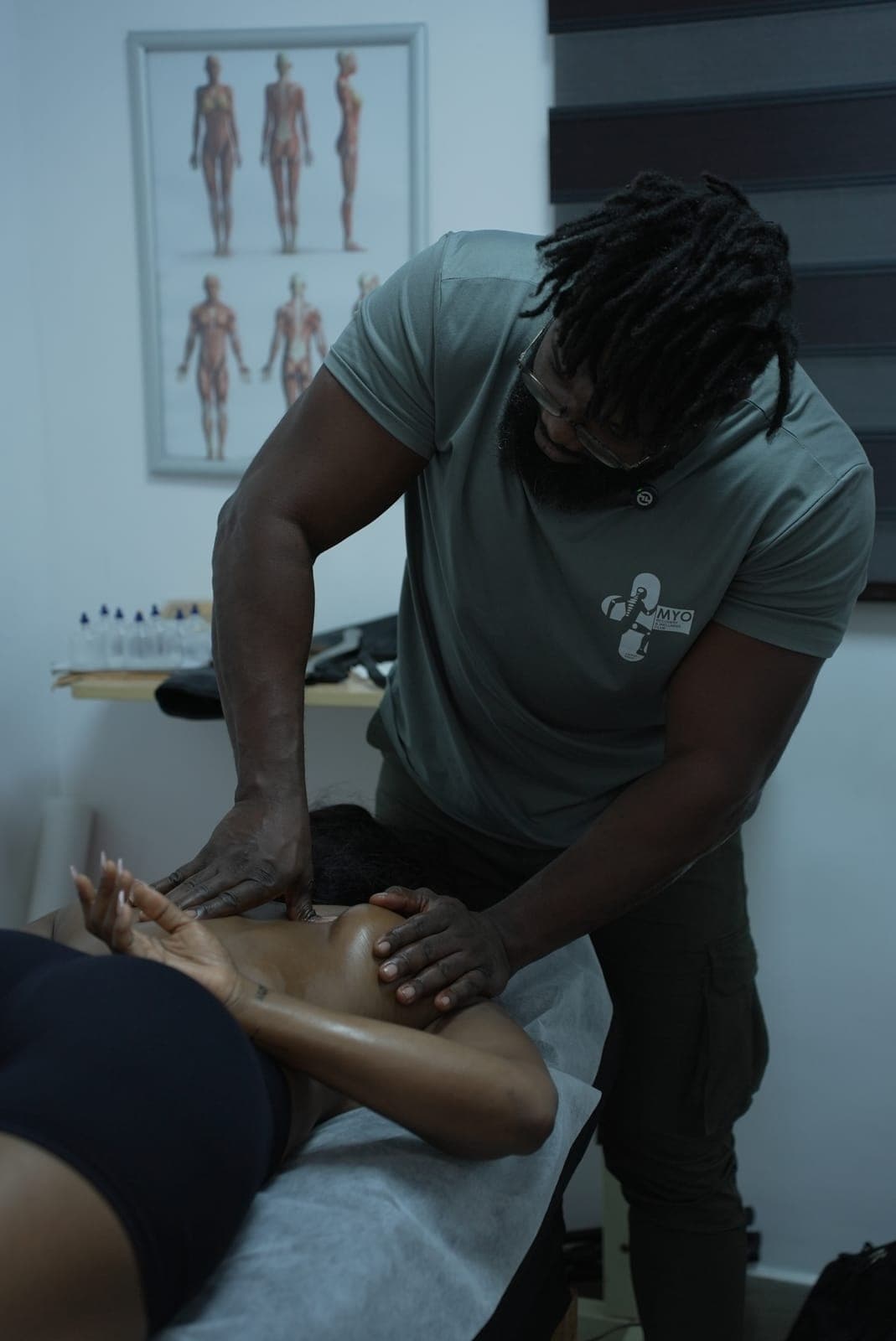 A MYO therapist in the MYO shirt working on a client's shoulder blade in the treatment room, an anatomy chart on the wall behind