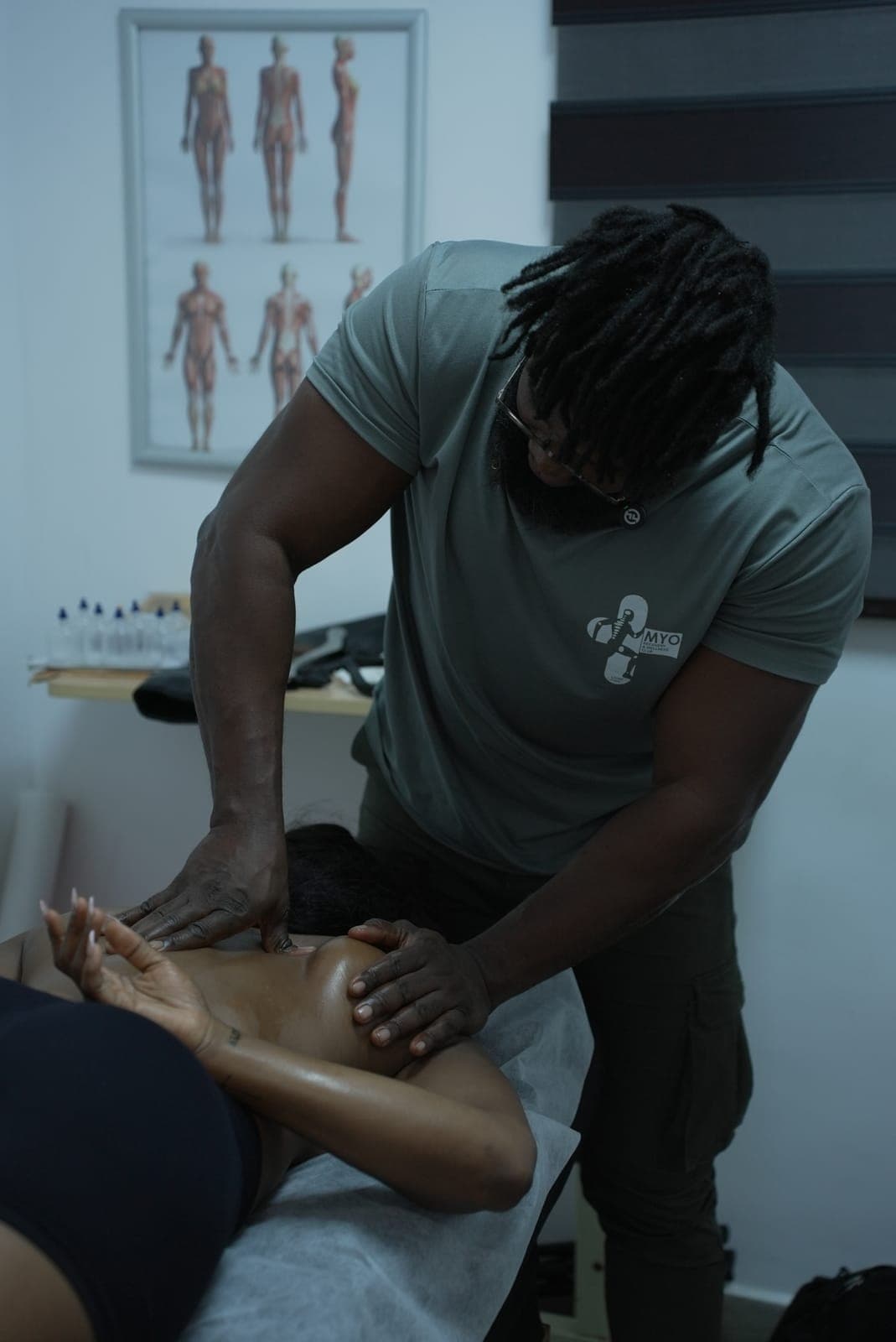 A MYO therapist in the MYO shirt pressing into a client's shoulder blade, the anatomy chart on the wall behind